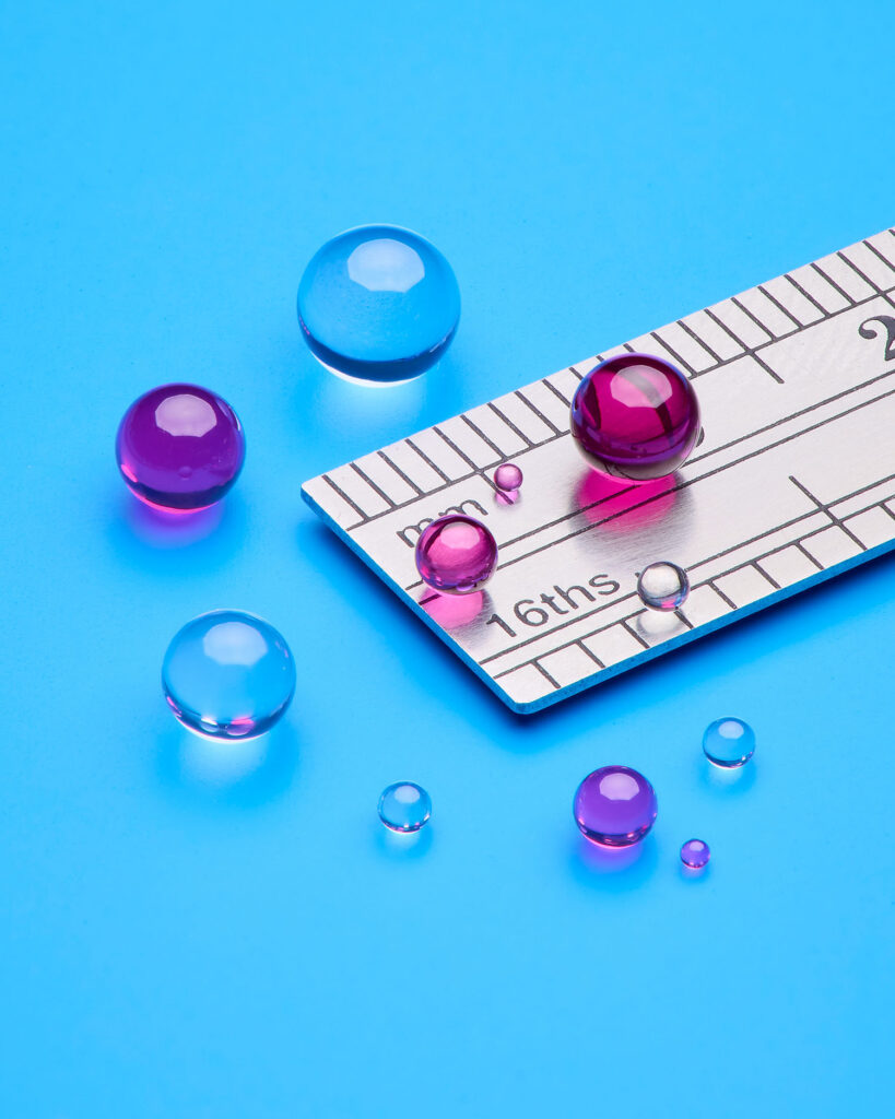 Tiny sapphire and ruby spheres sit on top of the edge of a silver metal ruler on a blue table.